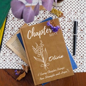Puede incluir: Un libro de tapa dura marrón con el texto Chapter SIXTY, el nombre Olivia y la frase Every Chapter Brings New Strength and Hope impresos en blanco. Un dibujo lineal blanco de un ramo de lirios de los valles está centrado en la portada.