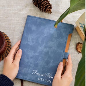 May include: A blue notebook with a textured, mottled finish featuring the white printed text "David Fin" and "MEd 2026" on the front cover. The notebook includes a blue elastic band closure and a brown ribbon bookmark extending from the bottom edge.