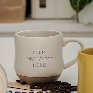 Puede incluir: Una taza de cerámica color crema con acabado moteado y asa, que presenta el texto en relieve YOUR TEXT/LOGO HERE en la parte frontal. La base de la taza es de un tono marrón que contrasta, rodeada de granos de café tostados oscuros.