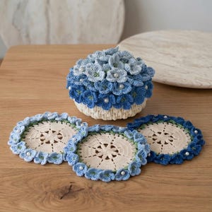 May include: A round, cream-colored crocheted container with a lid covered in blue flowers and small white pearls, accompanied by three matching round crocheted coasters featuring cream centers and blue floral borders with pearls.