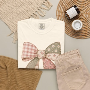 May include: A cream-colored short-sleeve t-shirt featuring a large, central graphic of a bow. The bow is composed of patchwork-style segments in muted pink and sage green, displaying patterns of gingham, small white daisies, and tiny floral prints.
