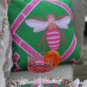May include: A pink and green patterned pillow with a pink and white bee design. A pink cocktail glass with a yellow and white rim sits on a silver tray with an orange slice garnish.