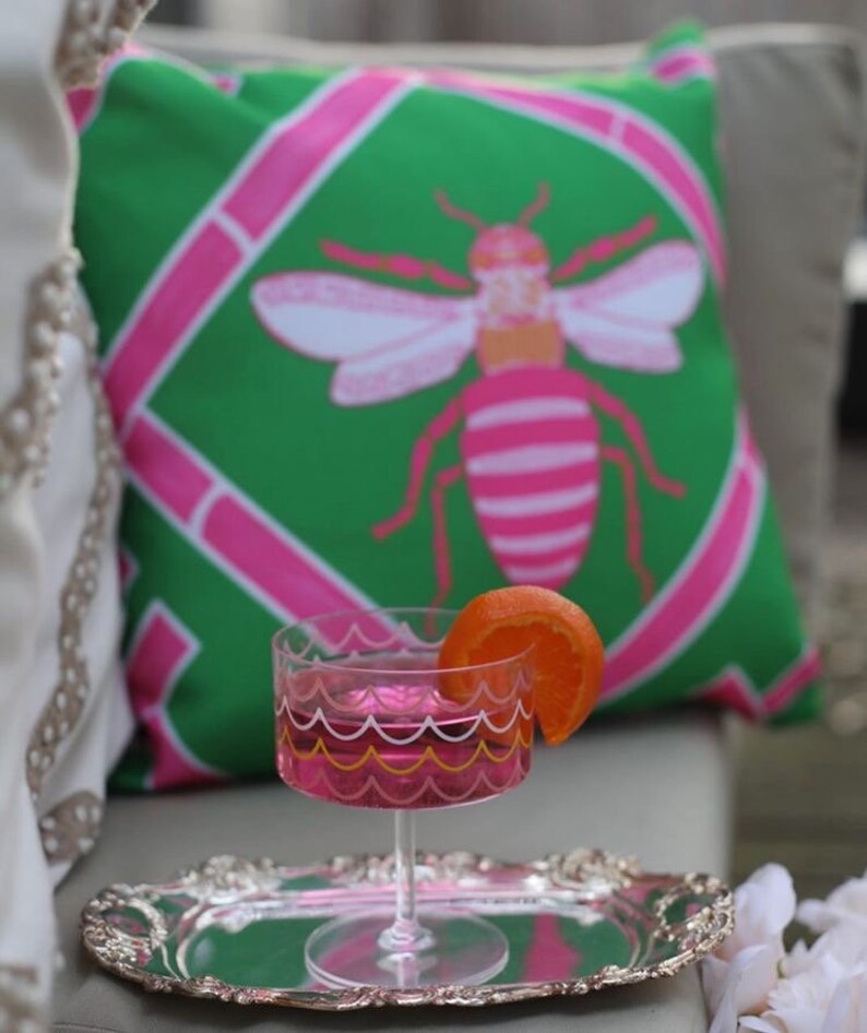 May include: A pink and green patterned pillow with a pink and white bee design. A pink cocktail glass with a yellow and white rim sits on a silver tray with an orange slice garnish.