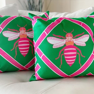 May include: Two green and pink throw pillows with a bee design. The bees are pink and white with orange accents. The pillows have a geometric pattern.