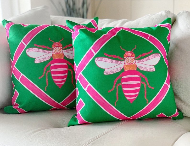May include: Two green and pink throw pillows with a bee design. The bees are pink and white with orange accents. The pillows have a geometric pattern.