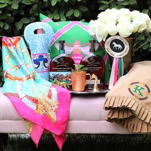 May include: A pink and green patterned pillow with a bee design, two bottles of Woodford Reserve bourbon, a copper mug, a brown blanket with a green letter K monogram, and a white floral arrangement in a brown ceramic vase with a horse-shaped ribbon.