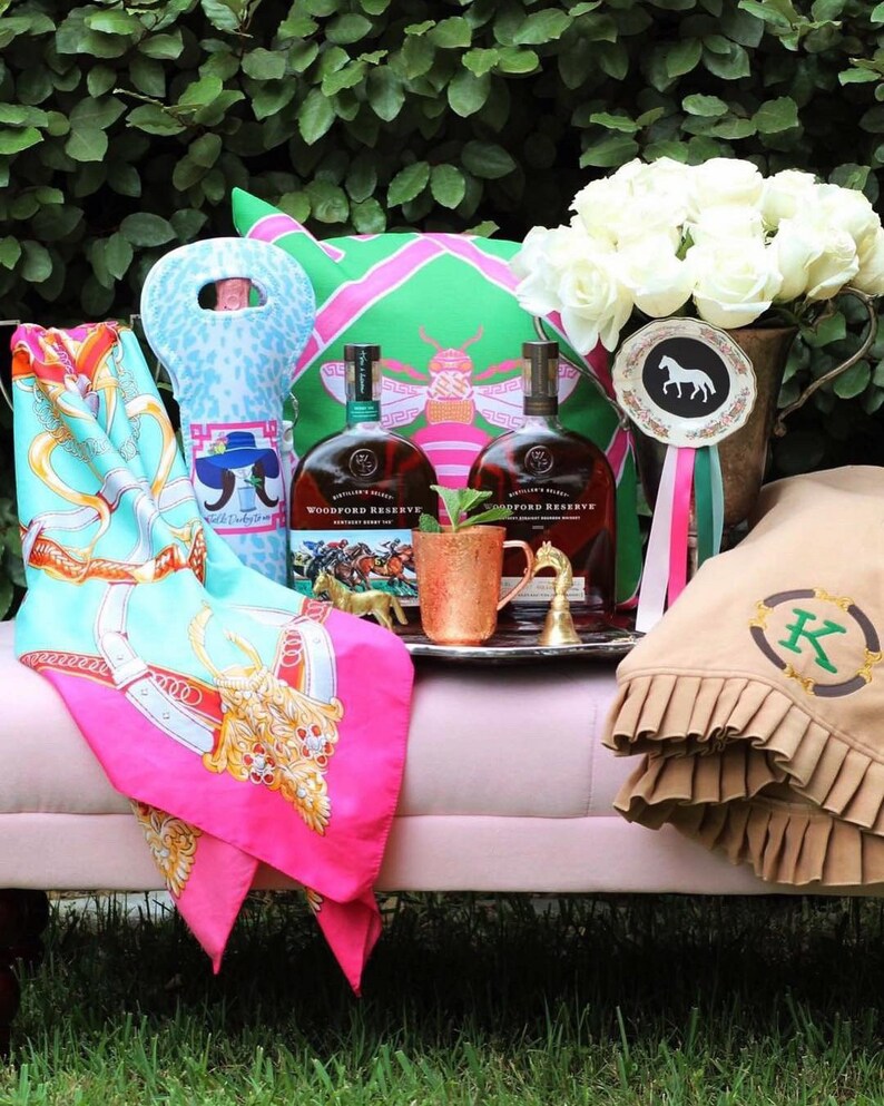May include: A pink and green patterned pillow with a bee design, two bottles of Woodford Reserve bourbon, a copper mug, a brown blanket with a green letter K monogram, and a white floral arrangement in a brown ceramic vase with a horse-shaped ribbon.
