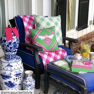 May include: A blue and white porcelain garden stool with a pink ribbon on top. The stool is next to a blue chair with a pink and white gingham throw and green and white gingham pillows. A green pillow with a pink bee design is on the chair. The chair is on a porch with a blue ottoman and a book on top.