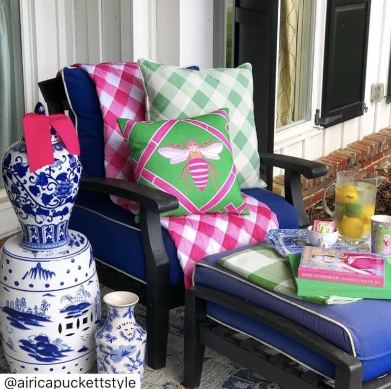 May include: A blue and white porcelain garden stool with a pink ribbon on top. The stool is next to a blue chair with a pink and white gingham throw and green and white gingham pillows. A green pillow with a pink bee design is on the chair. The chair is on a porch with a blue ottoman and a book on top.
