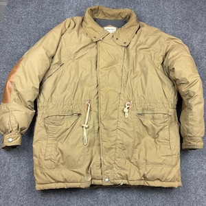 May include: A tan puffer jacket featuring a high collar, a front zip closure with a snap-button placket, and two large front pockets. The waist includes an adjustable drawstring with light-coloured toggles, and the sleeves have brown patches.