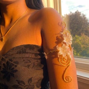 Puede incluir: Un brazalete de alambre en tono dorado con delicados detalles de flores blancas translúcidas y complejos diseños en espiral. La pieza está construida con alambre de metal fino y flexible formado en patrones orgánicos que envuelven la parte superior del brazo.