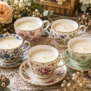 May include: Five tea cup candles with white wax and central wicks, each featuring distinct floral patterns and gold-trimmed edges. The collection includes cups with blue, pink, and green botanical designs, some paired with matching saucers.