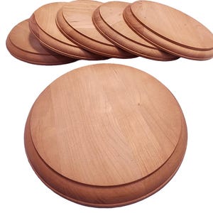 May include: A collection of unfinished, round wooden bases with a beveled edge. The bases are light brown in color and appear to be made of a hardwood. The image shows several bases stacked together, with one base in the foreground.