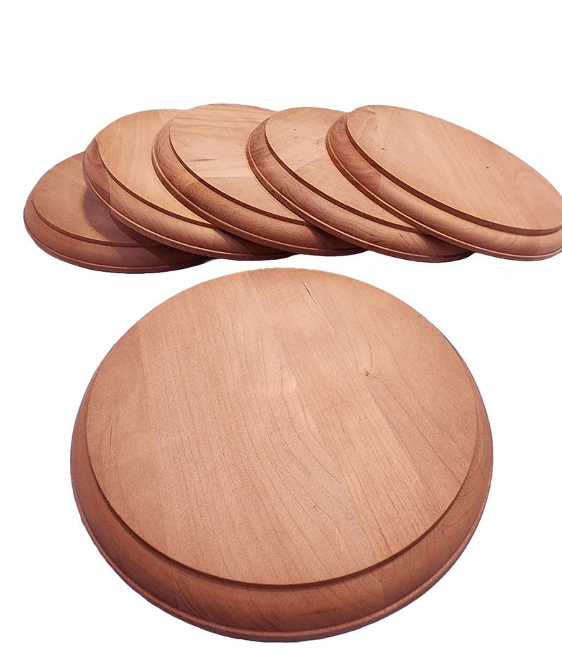 May include: A collection of unfinished, round wooden bases with a beveled edge. The bases are light brown in color and appear to be made of a hardwood. The image shows several bases stacked together, with one base in the foreground.