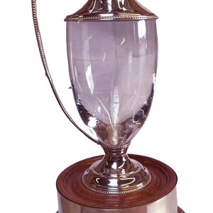 May include: A silver and glass pitcher with a curved handle and a decorative rim. The pitcher sits on a wooden base with a silver band. The design is elegant and classic, suitable for serving beverages.