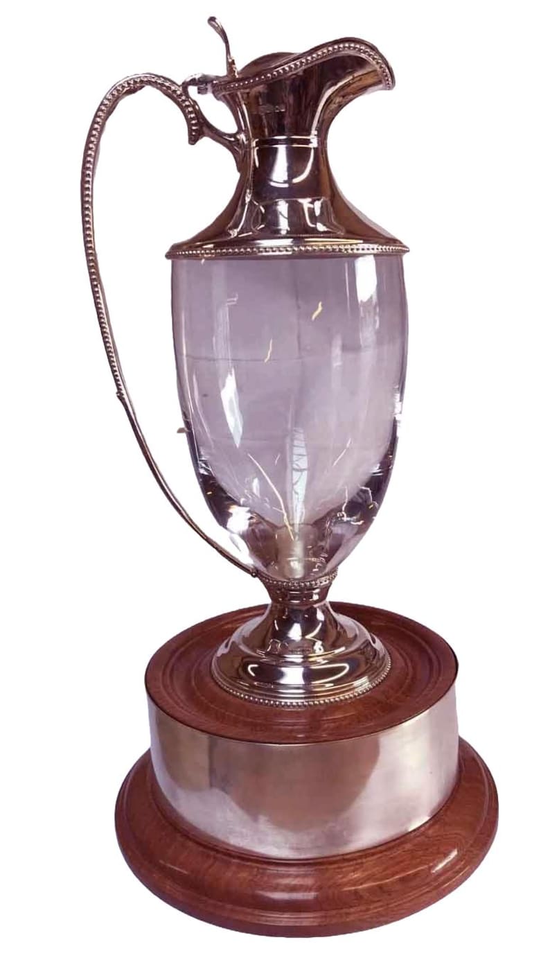 May include: A silver and glass pitcher with a curved handle and a decorative rim. The pitcher sits on a wooden base with a silver band. The design is elegant and classic, suitable for serving beverages.