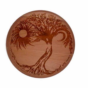 May include: Round wooden decorative piece with a carved tree design. The design incorporates a sun and moon, with intricate details in the branches and roots. The wood has a warm, natural tone.