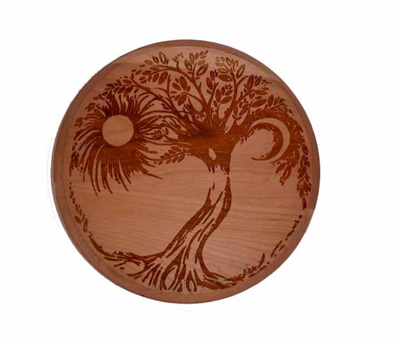 May include: Round wooden decorative piece with a carved tree design. The design incorporates a sun and moon, with intricate details in the branches and roots. The wood has a warm, natural tone.