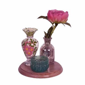 May include: A decorative arrangement featuring a pink peony in a small glass vase, a teal candle holder, and a floral-painted vase. The items are displayed on a round, wooden tray, creating a charming centerpiece.
