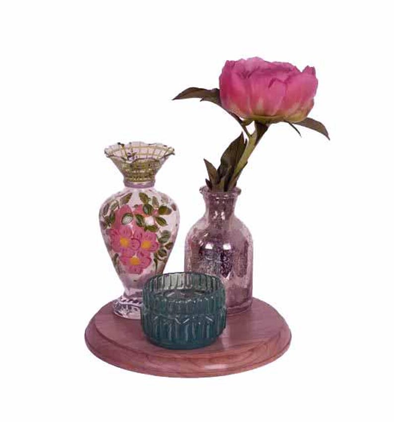 May include: A decorative arrangement featuring a pink peony in a small glass vase, a teal candle holder, and a floral-painted vase. The items are displayed on a round, wooden tray, creating a charming centerpiece.