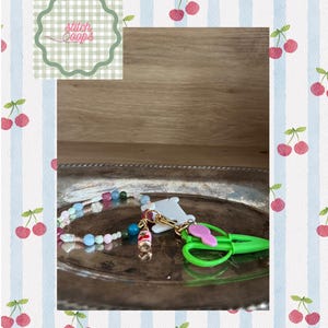 Könnte beinhalten: Ein Perlen-Accessoire bestehend aus einer Schnur aus bunten Glasperlen, einem kleinen Kirsch-Anhänger, einer weißen Kunststoff-Garnspule und einer hellgrünen Miniatur-Schere, befestigt mit einem goldfarbenen Karabinerverschluss.
