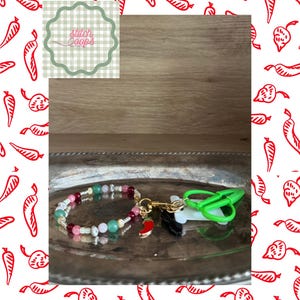 May include: A beaded accessory featuring a circular strand of multi-coloured beads in shades of pink, green, and white, accented with silver-toned spacers. A small red chilli pepper charm hangs from a gold-toned clasp attached to a green plastic scissor tool.