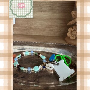 May include: A beaded accessory featuring light blue, turquoise, and dark brown beads, a white cowrie shell charm, a small blue heart charm, and a white plastic thread spool attached to a green plastic scissor-shaped charm on a metal ring.