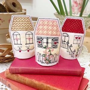 Peut inclure: Trois ornements brodés en forme de maison, avec différents motifs de toit et détails tels que fenêtres, portes et motifs floraux. Ils reposent sur une pile de livres rouges, créant une esthétique charmante et artisanale.