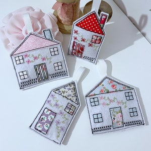 May include: Four handmade felt house decorations with different patterned fabric roofs. Each house has a white base with black embroidered details, including windows, doors, and a fence. The houses are hanging from white ribbon loops.