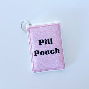 May include: A pink fabric pill pouch with a white floral pattern and black embroidered text that says "Pill Pouch". The pouch has a silver key ring attached.