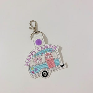 May include: A white keychain featuring a stitched camper design. The camper is light blue and pink with the words "HAPPY CAMPER" in purple. The keychain has a silver clasp and a purple button.