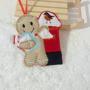 May include: A festive felt Christmas decoration. It features a gingerbread person holding letters, alongside a red postbox with a bird. The ornament has a red hanging ribbon and is set on a white, textured surface.