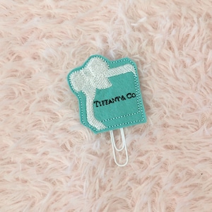 May include: A turquoise blue felt gift box with a white bow and the text "Tiffany & Co." embroidered on the front. The box is attached to a silver paper clip.