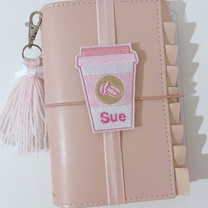 May include: A pink leather-bound journal with a pink elastic closure and a pink and white coffee cup patch with the name "Sue" embroidered on it. The journal has a pink tassel attached to it.