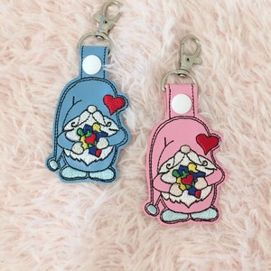 May include: Two gnome keyrings, one light blue and one pink, each with a red heart and holding a colourful bouquet. The keyrings have silver-tone clasps and are on a pink, fluffy surface.