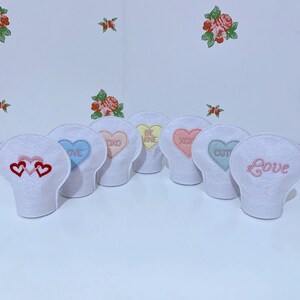Peut inclure: Six contenants en forme de cœur en feutre blanc avec différents messages de la Saint-Valentin brodés dessus. Les messages sont "Love", "Be Mine", "XOX", "Cutie", "Love", et "XOXO".