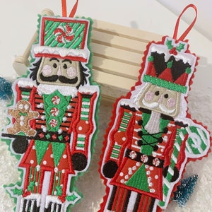 May include: Two felt Christmas decorations shaped like nutcrackers. One is red and green with a gingerbread man in its hand. The other is red, green, and black with a candy cane in its hand.
