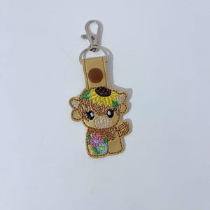 May include: A brown and tan cow keyring with a sunflower on its head and flowers in its hooves. The keyring is made of fabric and has a silver metal ring.
