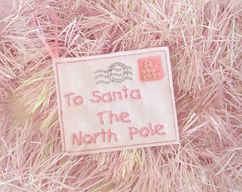 Könnte beinhalten: Ein weißer Filzbriefumschlag mit rosa Nähten und einem rosa "Elf Post" Stempel. Der Umschlag trägt die Aufschrift "To Santa The North Pole" in rosa Nähten.
