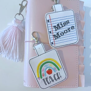May include: Two white fabric keychains with embroidered designs. One keychain has a rainbow with pencils and an apple, and the text "Teach!" The other keychain has a notebook with the text "Miss Moore".