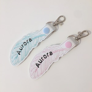 May include: Two feather-shaped keychains, one light blue and one pink, embroidered with the name "Aurora" in black. Each keychain has a white leather backing, a snap, and a silver-coloured clasp.
