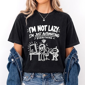 Może przedstawiać: Czarny t-shirt z krótkim rękawem z białym napisem graficznym o treści I'M NOT LAZY I'M JUST AUTOMATING EVERYTHING. Poniżej tekstu znajduje się rysunek liniowy zmęczonej osoby przy biurku komputerowym obok małego robota z napisem AI.