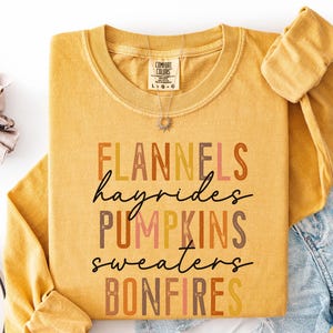 May include: A mustard yellow long-sleeve crewneck sweatshirt featuring the text FLANNELS, hayrides, PUMPKINS, sweaters, and BONFIRES in various earth-toned colors. The garment has a Comfort Colors brand label at the inner neckline.