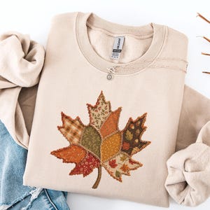 May include: A beige crewneck sweatshirt featuring a large, central maple leaf graphic composed of various patchwork-style fabric patterns in shades of orange, brown, green, and cream, with visible stitched detailing around the leaf edges.
