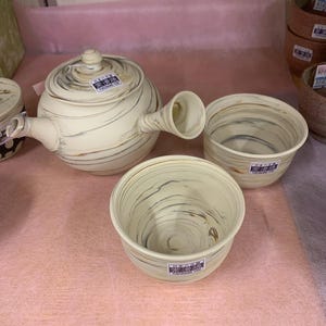 以下が含まれることがあります： サイドハンドル付きの陶器製急須と、お揃いの湯呑み2客のセット。全体がクリーム色をベースに、灰色と茶色のマーブル模様が施されています。各アイテムには、日本語のテキストと「TOKONAME-YAKI」と書かれた小さな長方形のステッカーが貼られています。