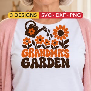 Puede incluir: Una camiseta blanca con un gráfico de una regadera marrón y flores naranjas con el texto 3 DESIGNS SVG DXF PNG y GRANDMA'S GARDEN en letras marrones y naranjas. El diseño incluye un girasol grande y flores más pequeñas.