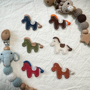 Peut inclure: Six écussons brodés en forme de cheval en bleu, marron, vert olive, crème, rouge et brun orangé, chacun avec une selle et une queue. Deux attaches-sucettes en crochet avec des perles en bois et des étoiles sont placées aux coins.