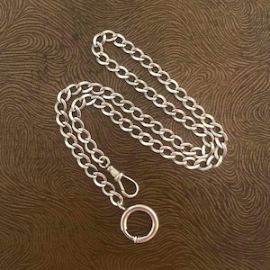 May include: A silver-colored chain with a lobster clasp and a circular clasp. The chain is made of oval links and is displayed on a brown surface.