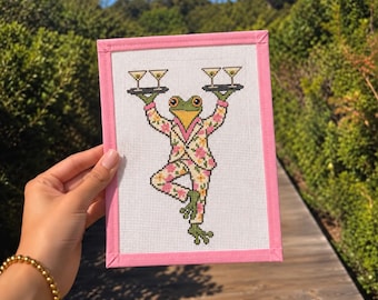 May include: A rectangular needlepoint canvas featuring a cross-stitched frog wearing a floral suit, balancing two trays with martini glasses on its hands. The canvas is framed with a light pink fabric border on a white mesh background.