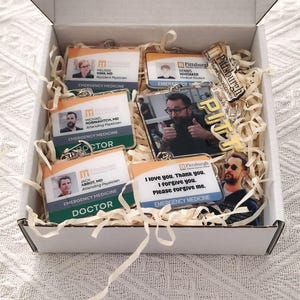 May include: A white cardboard box filled with shredded paper and several rectangular acrylic keychains. The keychains feature photos of individuals, medical identification text, and the word PITT in gold lettering. One keychain displays the text I love you. Thank you. I forgive you. Please forgive me.